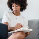 woman sitting on a couch writing in a notebook and looking at a laptop screen