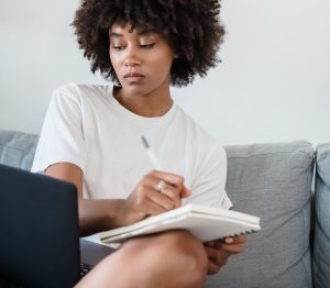woman sitting on a couch writing in a notebook and looking at a laptop screen