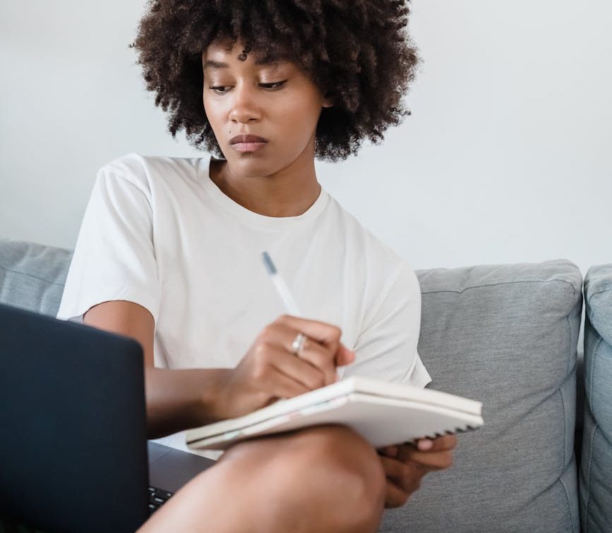 woman sitting on a couch writing in a notebook and looking at a laptop screen