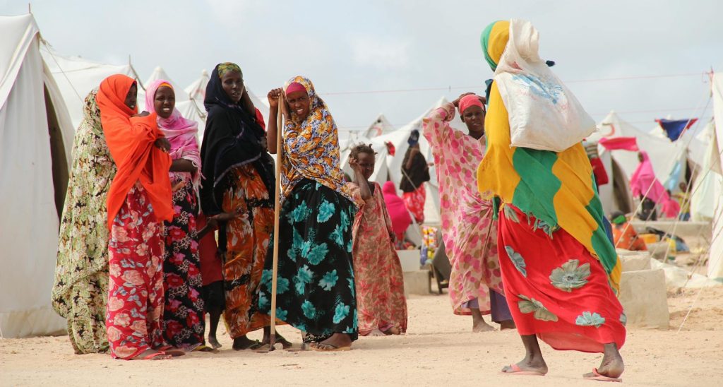 a group of women in colorful gowns standing between tents in a village