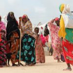a group of women in colorful gowns standing between tents in a village